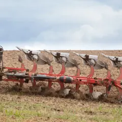 Czerwony pług z wieloma skibami pracuje na polu, przygotowując ziemię do siewu.