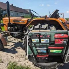 Pomarańczowy Ursus C-330, klasyczny ciągnik rolniczy, stoi na podwórku. W rogu widać półkę z akumulatorami ZAP.