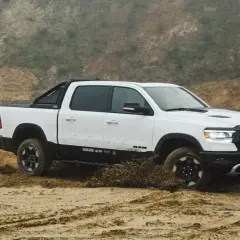 Biały Dodge Ram 1500 z czarnymi dodatkami pokonuje piaszczysty teren, wzbijając błoto.