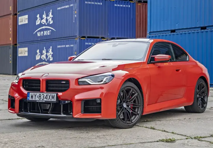 Pomarańczowe BMW M2 Competition z czarnymi felgami stoi na tle niebieskich kontenerów.