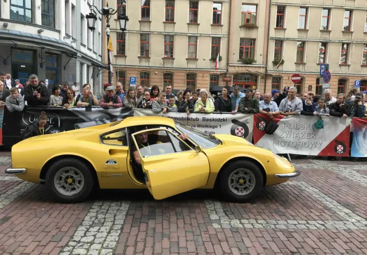 Żółte Ferrari Dino z otwartymi drzwiami, kierowca w środku, tłum widzów za barierką.