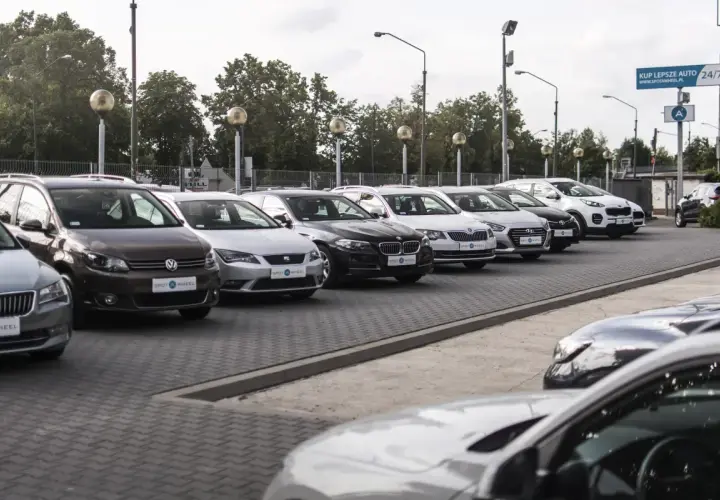Rząd samochodów na placu, w tym brązowy Volkswagen i biały Kia Sportage. W tle drzewa i słupy oświetleniowe. To idealne miejsce, by znaleźć swój wymarzony kinto one.