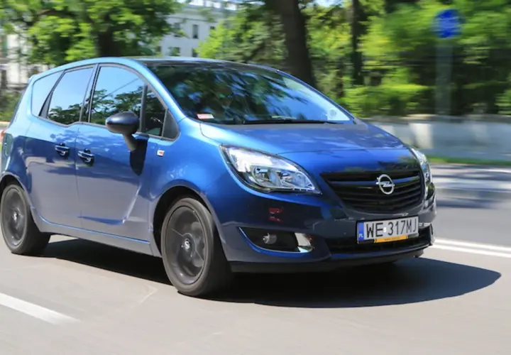 Niebieski Opel Meriva 1.4 benzyna w ruchu miejskim, z rozmazanym tłem drzew i budynków.
