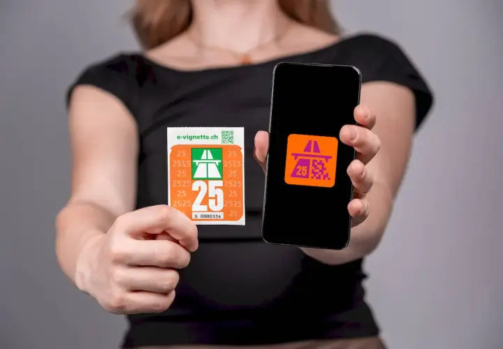 Ręce trzymające naklejkę i smartfon z aplikacją e-winieta szwajcaria. Na naklejce i ekranie telefonu widnieje symbol autostrady i cyfra 25.