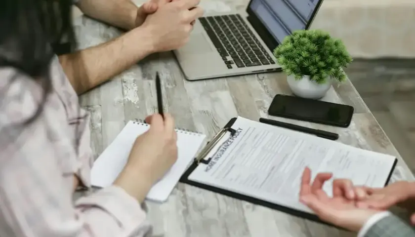 Osoby przy stole omawiają dokument "Home Insurance Policy". Kobieta notuje, mężczyzna patrzy na laptopa. Może to być związane z wefox wypowiedzenie oc.