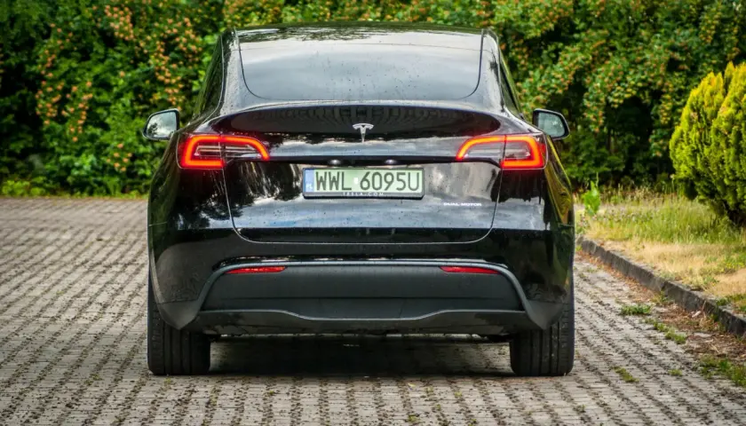 Czarna Tesla z napędem dual motor, z tablicą rejestracyjną WWL 6095U, zaparkowana na kostce brukowej wśród zieleni.