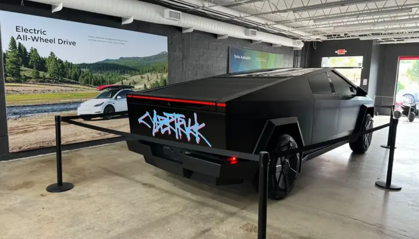 Czarny Tesla Cybertruck z neonowym napisem "CYBERTRUCK" na tyle. W tle reklama "Electric All-Wheel Drive". Cena tego futurystycznego pojazdu budzi zainteresowanie.