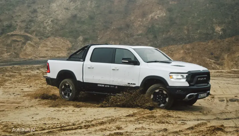 Biały Dodge Ram 1500 z czarnymi dodatkami pokonuje piaszczysty teren, wzbijając błoto.