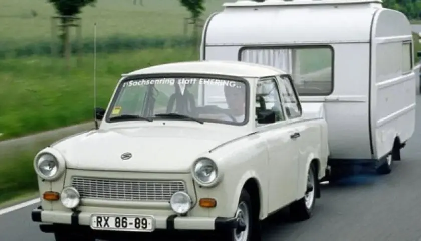 Biały trabant ciągnie przyczepę kempingową po drodze.