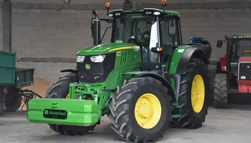 Zielony John Deere 6195M, z kierowcą w kabinie, stoi obok przyczepy. Czy to najlepsza marka ciągnika?