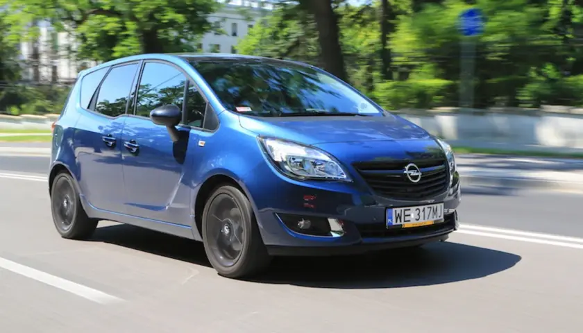 Niebieski Opel Meriva 1.4 benzyna w ruchu miejskim, z rozmazanym tłem drzew i budynków.