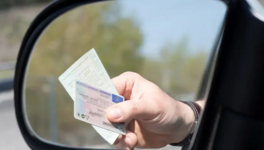 Dłoń trzyma prawo jazdy i dowód rejestracyjny. Kierowca pokazuje dokumenty, by uniknąć mandatu za brak prawa jazdy.