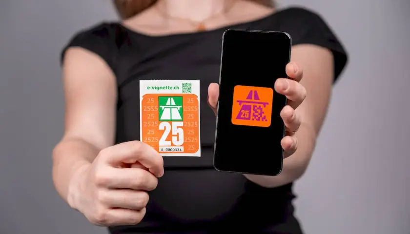 Ręce trzymające naklejkę i smartfon z aplikacją e-winieta szwajcaria. Na naklejce i ekranie telefonu widnieje symbol autostrady i cyfra 25.
