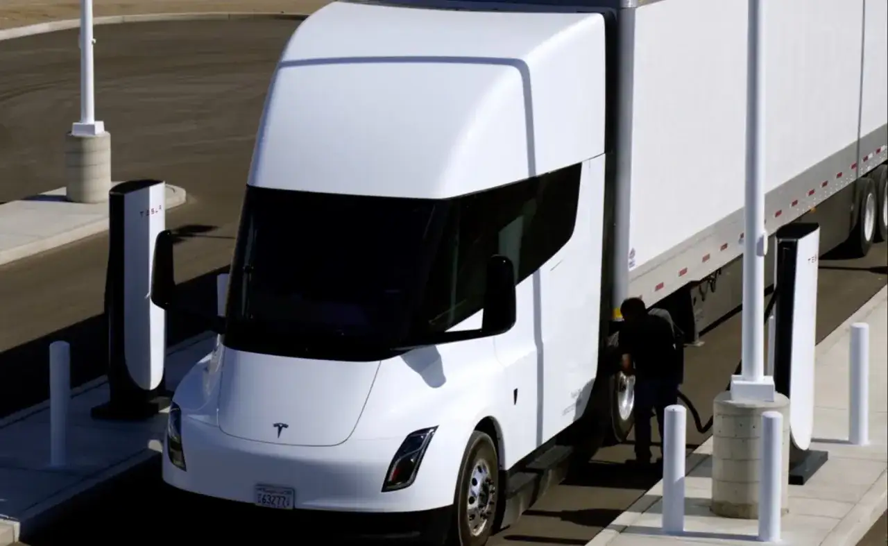 Biały Tesla Semi ładuje się na stacji. Kierowca podłącza kabel do ciężarówki.