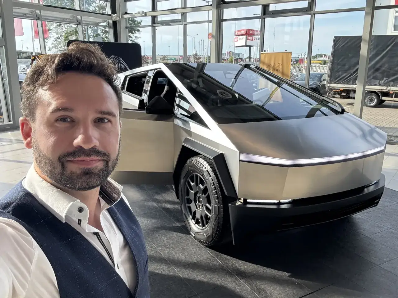 Mężczyzna w garniturze pozuje do selfie obok futurystycznego Cybertrucka w salonie.