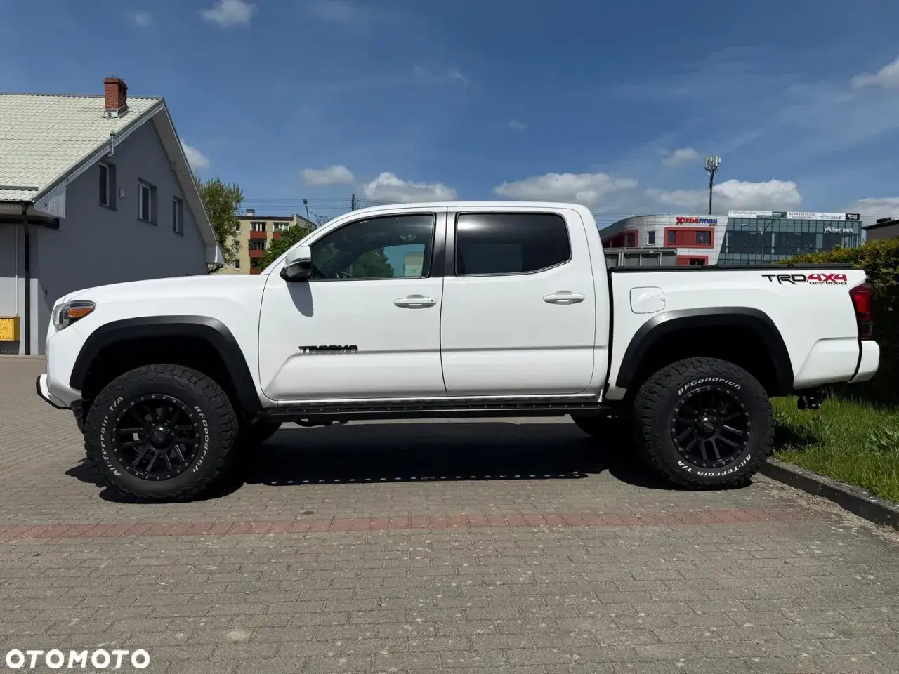 Biała Toyota Tacoma TRD Off-Road z czarnymi felgami i oponami BFGoodrich All-Terrain T/A, zaparkowana na chodniku.